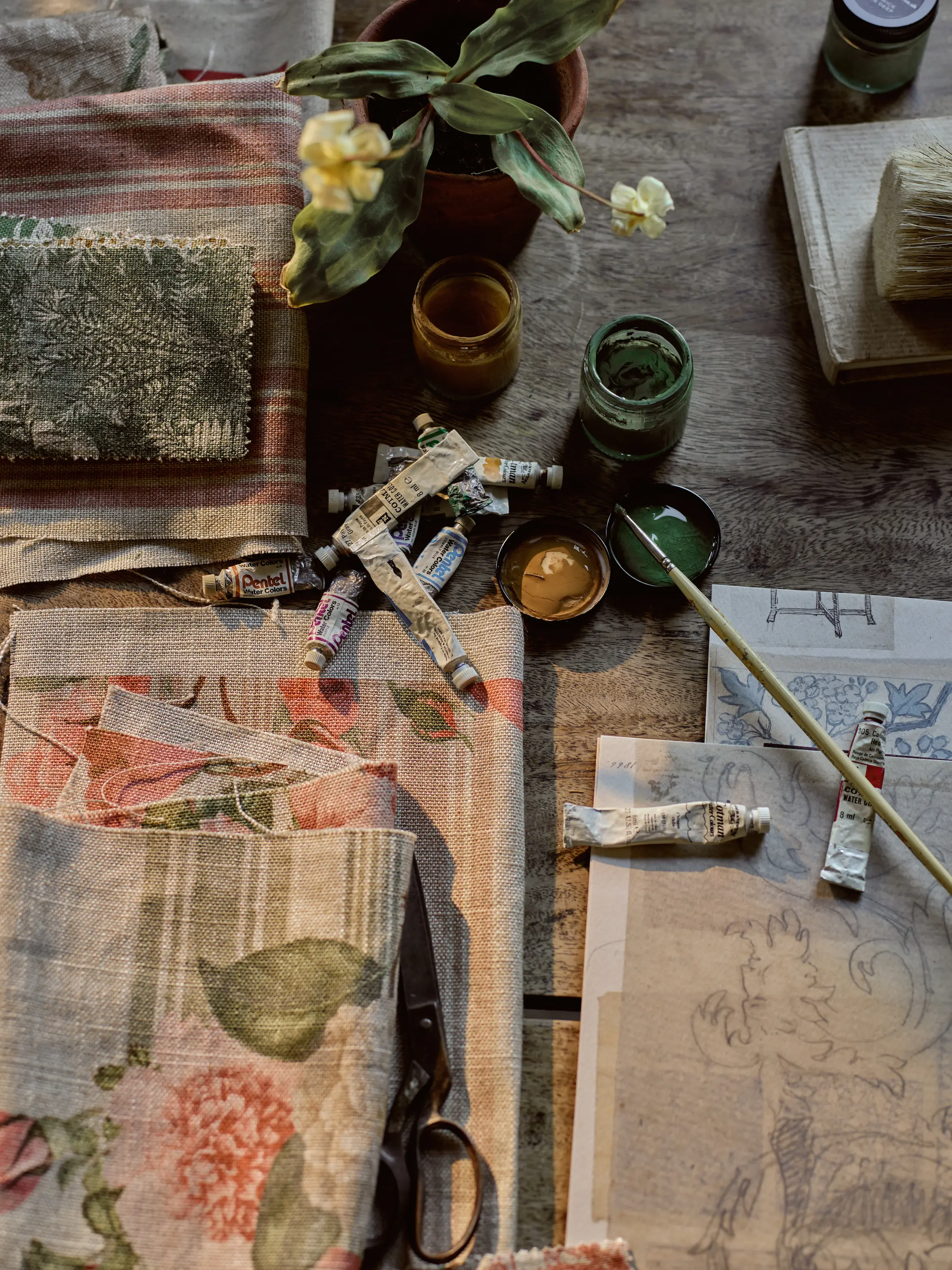 A wooden table with floral fabric swatches, paint tubes, jars of green and gold paint, paintbrushes, sketch papers with drawings, a pair of scissors, and a potted plant with yellow flowers.